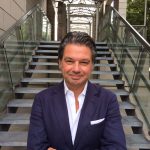 Ein Mann in einem marineblauen Anzug steht selbstbewusst mit verschränkten Armen vor einer modernen Außentreppe.