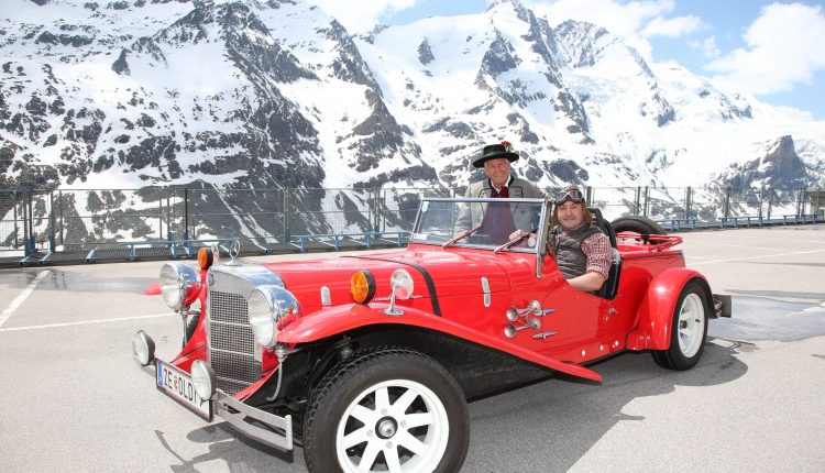 Zwei Personen in einem roten Oldtimer-Cabrio mit verschneiten Bergen im Hintergrund an einem sonnigen Tag.