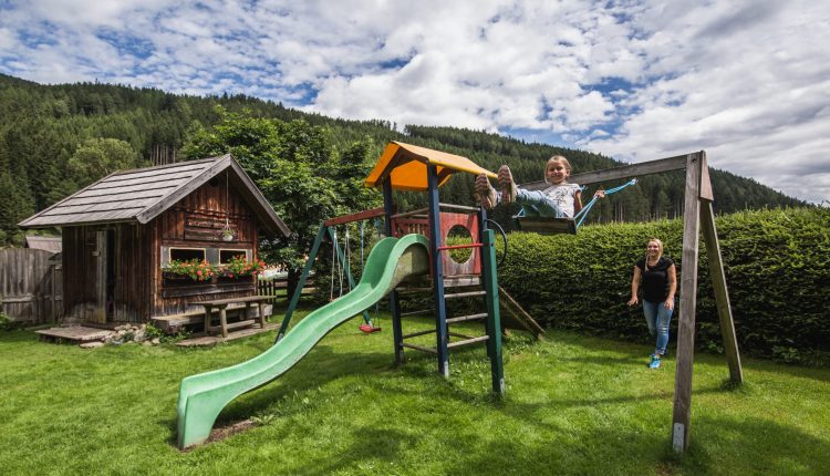 Ein Kind schaukelt neben einer Rutsche in einem grasbewachsenen Hof, im Hintergrund sind eine Frau und ein Holzhaus zu sehen.