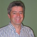 Ein Mann mit grauem Haar, der ein kariertes Hemd trägt, lächelt vor einem einfarbigen grünen Hintergrund.