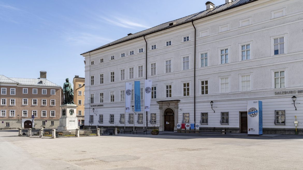 Großes weißes historisches Gebäude mit Fahnen und Statue auf einem ruhigen, offenen Platz an einem sonnigen Tag.