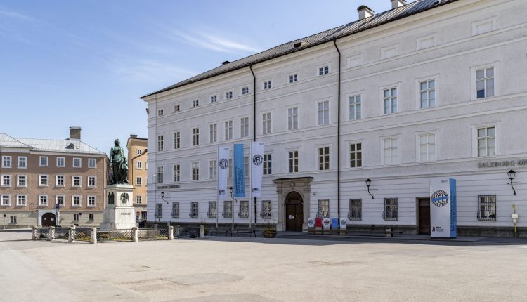 Großes weißes historisches Gebäude mit Fahnen und Statue auf einem ruhigen, offenen Platz an einem sonnigen Tag.