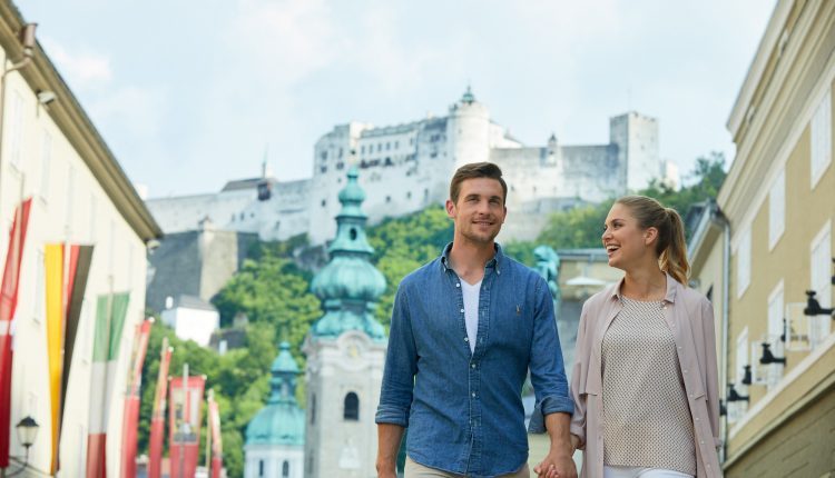 Ein lächelndes Paar hält sich an den Händen, während es in einer europäischen Stadt spazieren geht, mit einer Burg auf einem Hügel im Hintergrund.
