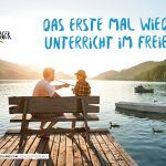 Zwei Menschen sitzen auf einem Steg an einem See und unterhalten sich, während im Hintergrund Berge, Boote und der Sonnenuntergang zu sehen sind.