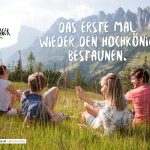 Vier junge Menschen sitzen auf einer Wiese mit Blick auf die Berge unter einem blauen Himmel, lächeln und unterhalten sich.