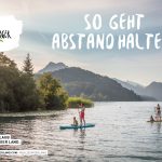 Zwei Personen paddeln und eine Person sitzt auf einem Paddelbrett auf einem See mit Bergen im Hintergrund.