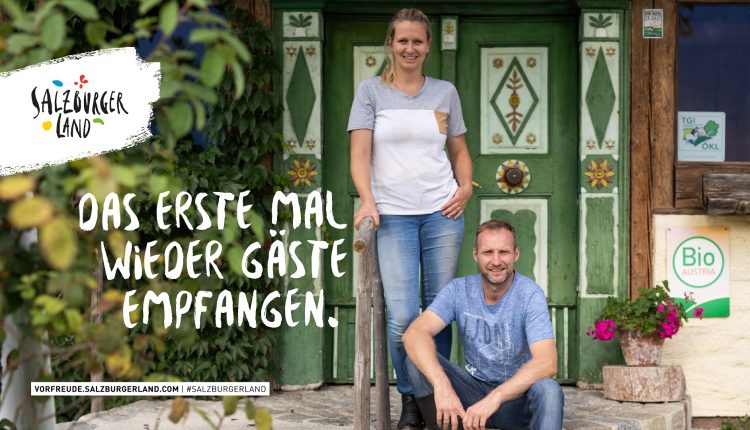 Ein lächelnder Mann und eine lächelnde Frau stehen vor einer rustikalen Holztür mit dem Text Das erste Mal wieder Gäste empfangen.