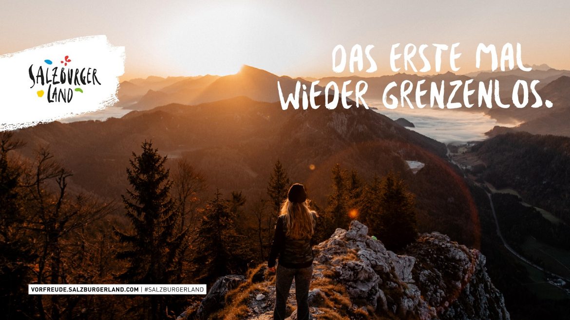 Eine Person steht bei Sonnenaufgang auf einem Berggipfel und blickt auf ein malerisches Tal und ferne Berge.