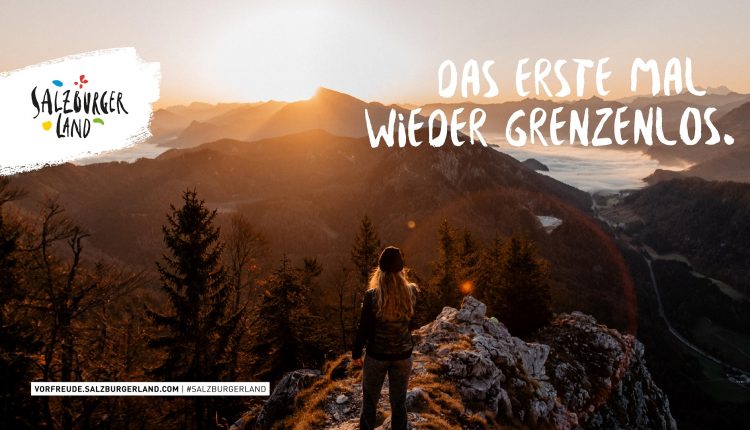 Eine Person steht bei Sonnenaufgang auf einem Berggipfel und blickt auf ein malerisches Tal und ferne Berge.