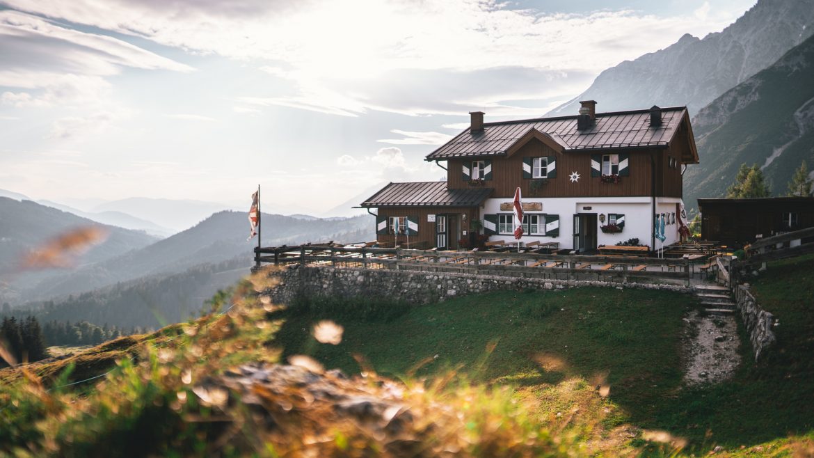 Eine hölzerne Berghütte liegt auf einer grasbewachsenen Anhöhe mit malerischen Bergen und einem strahlenden Himmel im Hintergrund.