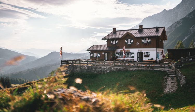 Eine hölzerne Berghütte liegt auf einer grasbewachsenen Anhöhe mit malerischen Bergen und einem strahlenden Himmel im Hintergrund.