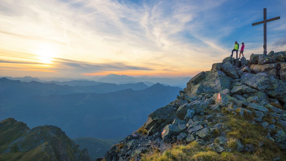 Zwei Wanderer stehen bei Sonnenuntergang an einem großen Kreuz auf einem felsigen Berggipfel und blicken auf ferne Hügel und Täler.