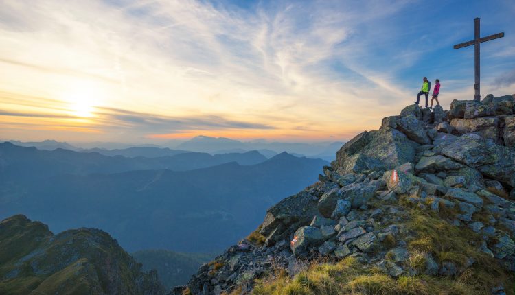 Zwei Wanderer stehen bei Sonnenuntergang an einem großen Kreuz auf einem felsigen Berggipfel und blicken auf ferne Hügel und Täler.