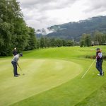 Drei Personen spielen Golf auf einem grünen Platz mit Bergen und Bäumen im Hintergrund unter bewölktem Himmel.