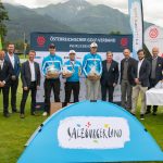 Eine Gruppe posiert auf einem Golfplatz mit Trophäen, Bannern und Bergen im Hintergrund.