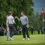 Zwei Golfer stoßen sich auf dem Grün die Fäuste, während andere zuschauen, mit Bäumen und Bergen im Hintergrund.