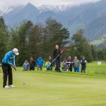 Ein Golfer spielt einen Putt auf dem Grün, während die Zuschauer zusehen, mit Bergen und Bäumen im Hintergrund.