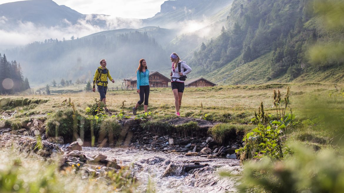 Drei Personen wandern entlang eines Baches in einem grasbewachsenen Bergtal mit Hütten im österreichischen Stil und Nebel im Hintergrund.