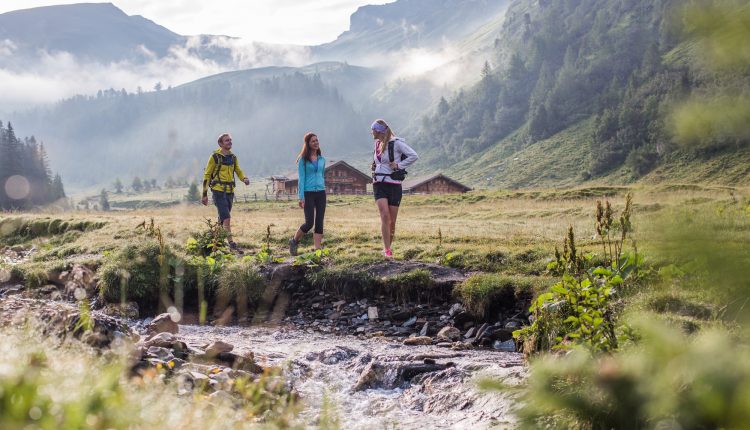 Drei Personen wandern entlang eines Baches in einem grasbewachsenen Bergtal mit Hütten im österreichischen Stil und Nebel im Hintergrund.