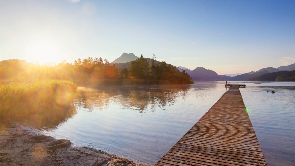 Der hölzerne Steg führt in einen ruhigen See bei Sonnenaufgang, mit Bäumen und Bergen in der Ferne.