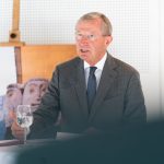 Ein älterer Mann im Anzug spricht an einem Tisch mit einem Glas Wasser und einem Plakat im Hintergrund.