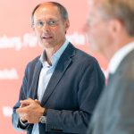 Ein Mann in einem blauen Blazer spricht, während ein anderer Mann in einem Anzug unscharf im Vordergrund steht.