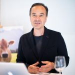 Ein Mann in einem dunklen Blazer spricht an einem Schreibtisch mit einem Laptop, einem leeren Weinglas und einem Kuhposter im Hintergrund.