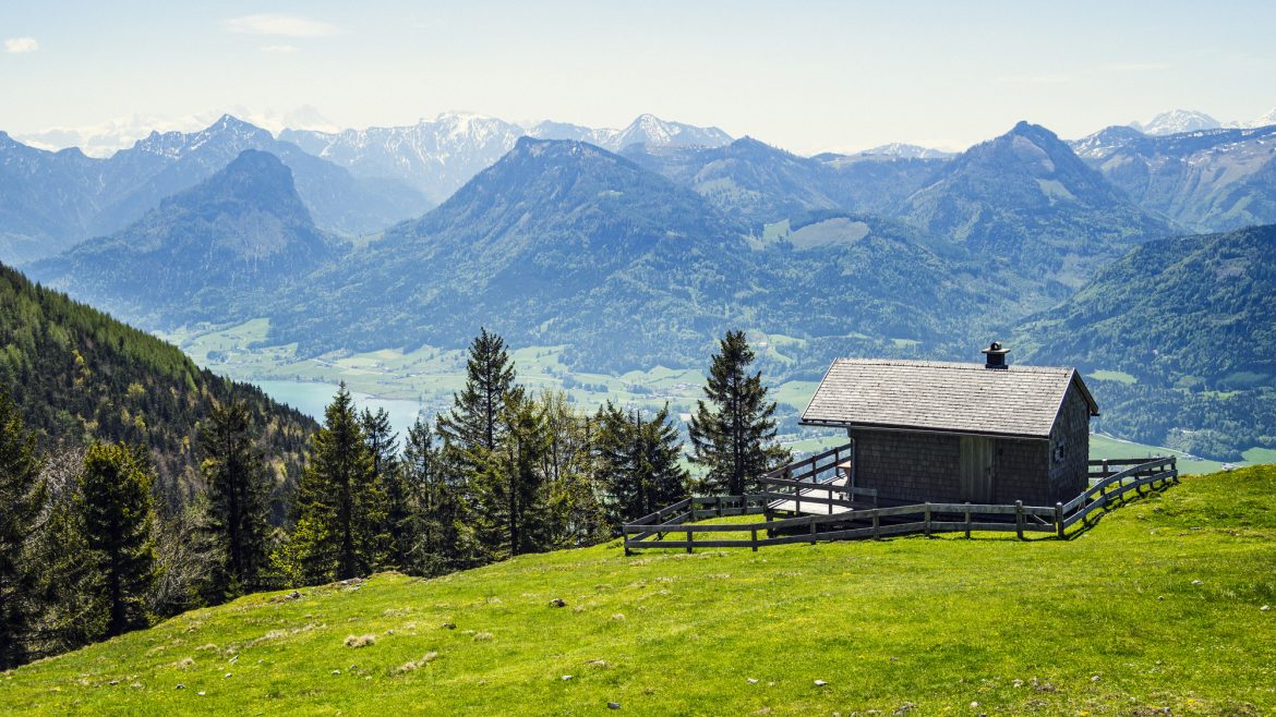 Eine kleine Hütte auf einem grasbewachsenen Hügel überblickt die fernen Berge und einen See unter einem klaren blauen Himmel.