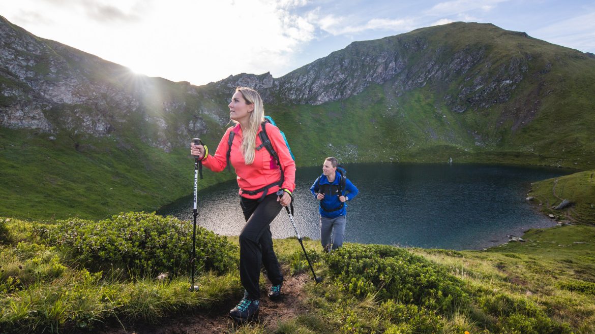 Zwei Personen wandern bergauf auf einem grasbewachsenen Bergpfad in der Nähe eines Sees mit Bergen im Hintergrund.