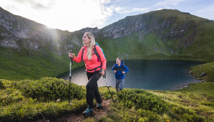 Zwei Personen wandern bergauf auf einem grasbewachsenen Bergpfad in der Nähe eines Sees mit Bergen im Hintergrund.
