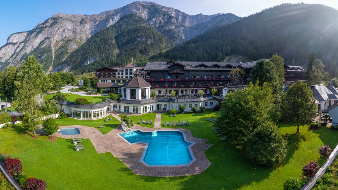 Ein Luxushotel mit Pool und Gärten, umgeben von bewaldeten Bergen und einem klaren Himmel.