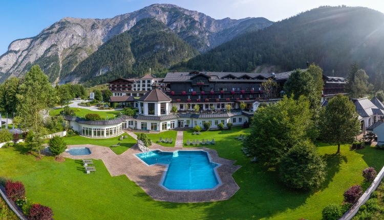 Ein Luxushotel mit Pool und Gärten, umgeben von bewaldeten Bergen und einem klaren Himmel.