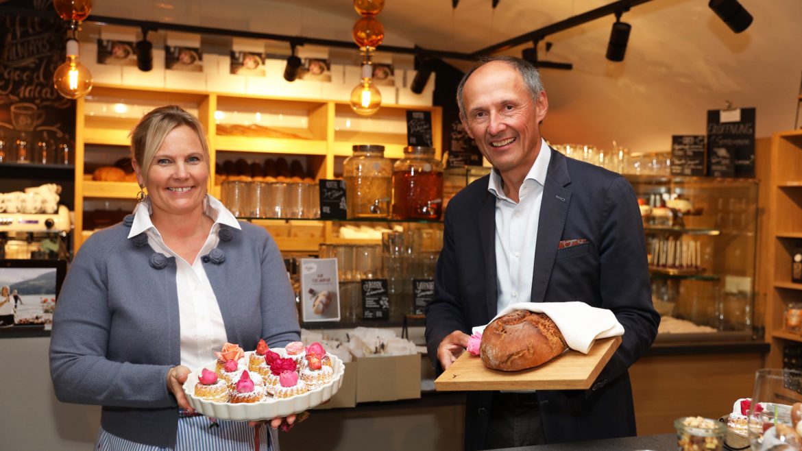 Eine lächelnde Frau hält ein Tablett mit Muffins in der Hand, während ein Mann in einer gemütlichen Bäckerei einen Laib Brot präsentiert.
