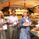 Vier lächelnde Menschen in einer Bäckerei, die Geschenke, Backwaren und einen Kuchen in der Hand halten und in einem gemütlichen, warm beleuchteten Laden stehen.