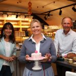 Drei lächelnde Menschen in einer Küche, eine Frau hält einen kleinen rosa Kuchen auf einem Ständer.