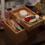 Holzkiste mit Baguettescheiben, Dip, Gurken, Kirschtomaten und Oliven auf einem dekorierten Tisch.