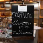 Ein Schild auf einem Tresen kündigt die Eröffnung des Cafés am 1. September um 8.30 Uhr an; im Hintergrund Regale.