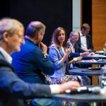 Fünf Personen sitzen an einem Podiumstisch, eine Frau spricht in ein Mikrofon, die anderen hören aufmerksam zu.
