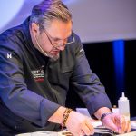 Ein Koch in schwarzer Uniform serviert an einem Tresen bei heller Beleuchtung sorgfältig das Essen.