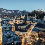 Luftaufnahme von Salzburg in der Abenddämmerung, mit beleuchteten Straßen, historischen Gebäuden und einer Festung auf einem Hügel.
