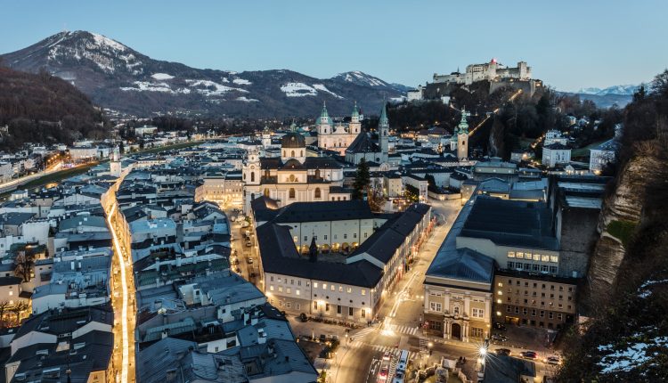 Luftaufnahme von Salzburg in der Abenddämmerung, mit beleuchteten Straßen, historischen Gebäuden und einer Festung auf einem Hügel.