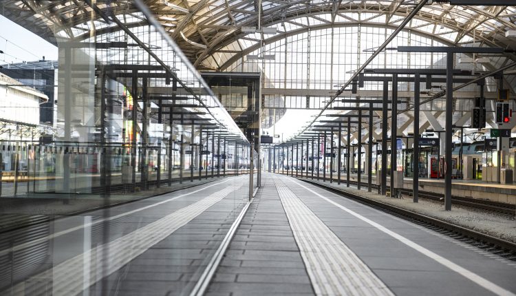 Leerer Bahnsteig mit Metallträgern, Glasdach und Gleisen, die sich in der Glaswand des Bahnhofs spiegeln.