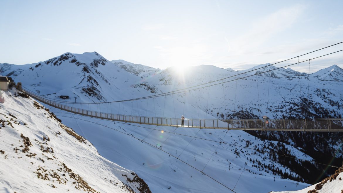 Eine Hängebrücke überquert bei Sonnenaufgang verschneite Berge, in deren Mitte eine Person läuft.