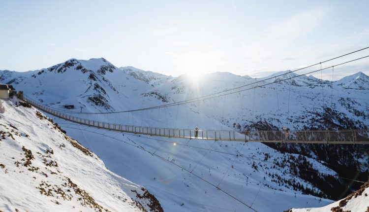 Eine Hängebrücke überquert bei Sonnenaufgang verschneite Berge, in deren Mitte eine Person läuft.