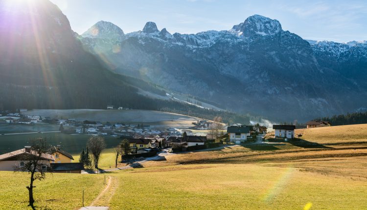 Ein sonnenbeschienenes Alpendorf mit Bergen im Hintergrund und grünen Feldern im Vordergund.