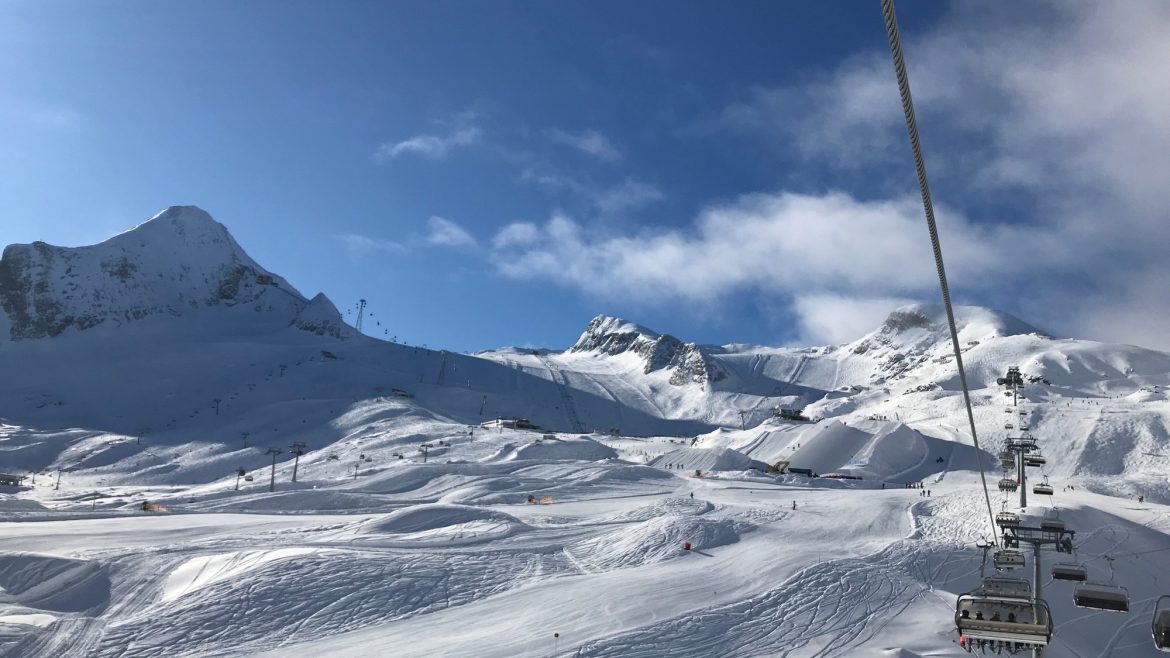 Skilift, der über verschneite Berge unter einem blauen Himmel mit Sonnenlicht und vereinzelten Wolken aufsteigt.