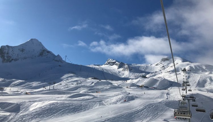 Skilift, der über verschneite Berge unter einem blauen Himmel mit Sonnenlicht und vereinzelten Wolken aufsteigt.