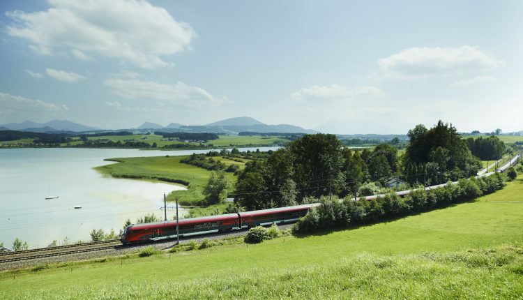 Ein roter Zug fährt an einem See entlang durch eine grüne Landschaft unter einem klaren blauen Himmel.