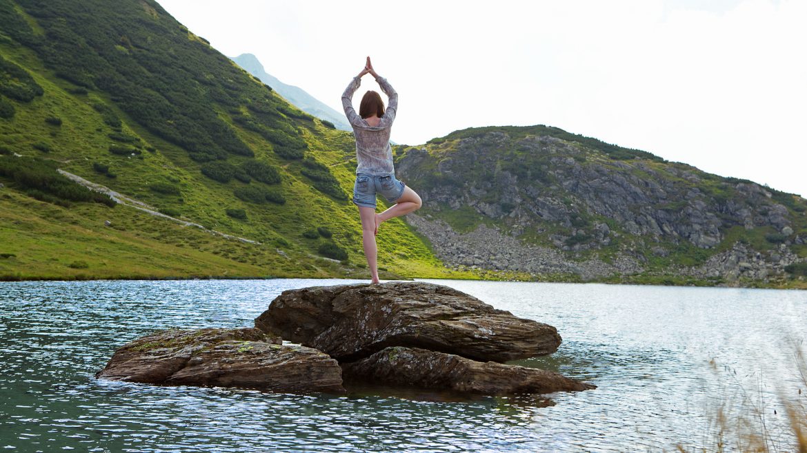 Person beim Yoga auf einem Felsen in einem See, umgeben von grünen Hügeln und hellem Tageslicht.