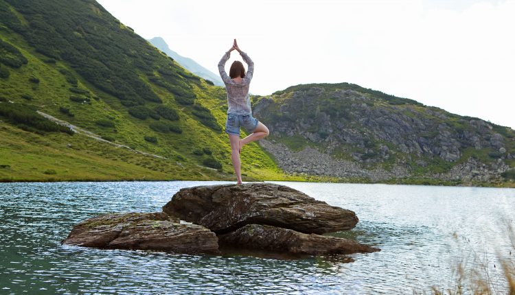 Person beim Yoga auf einem Felsen in einem See, umgeben von grünen Hügeln und hellem Tageslicht.